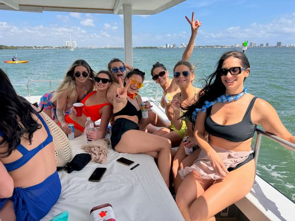 Group of friends in swimsuits on a sunny party boat in a blue bay, laughing and posing with a coastal city skyline and kayaker in the background.