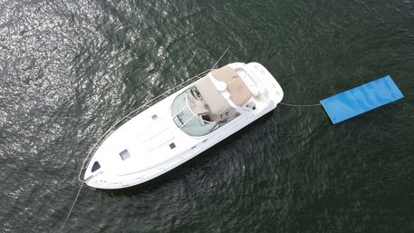 Aerial view of a white motor yacht anchored in sun-sparkled dark green water, tethered to a bright blue floating swim mat.