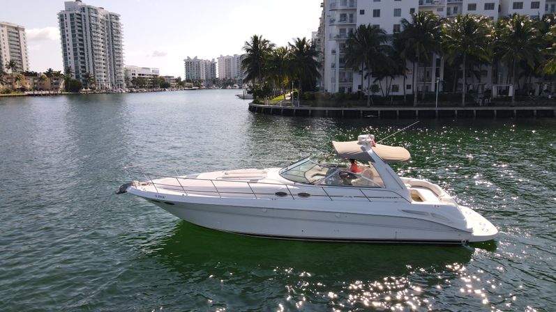 White motor yacht cruising a sunny waterfront canal with sparkling green water, palm trees and high-rise condominiums