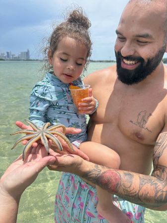 Smiling father holding his toddler at a sunny beach, both examining a large starfish in clear turquoise shallow water with a coastal skyline in the distance.