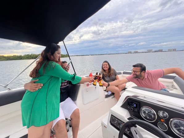 Group of friends on a motorboat cheering with beer bottles at sunset over calm bay waters and a distant coastal skyline