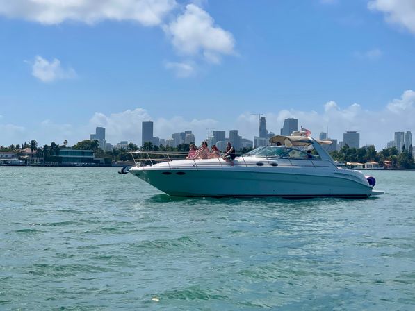 White cabin cruiser with people lounging on deck on turquoise bay, palm-lined shore and Miami skyline under a sunny blue sky.