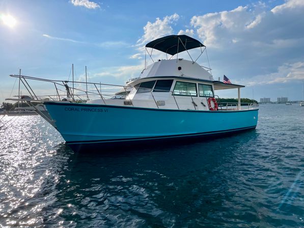 Turquoise-hulled motor yacht anchored in a sunny coastal harbor, sparkling blue water, shaded flybridge and American flag visible