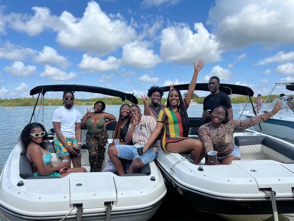 Group of friends enjoying a sunny boat party on two linked motorboats in calm blue water, smiling and waving under puffy clouds — summer waterfront outing.