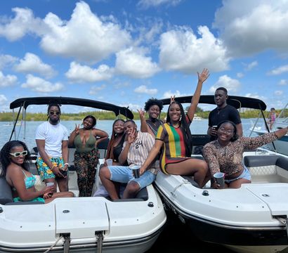 Group of friends enjoying a sunny boat party on two linked motorboats in calm blue water, smiling and waving under puffy clouds — summer waterfront outing.