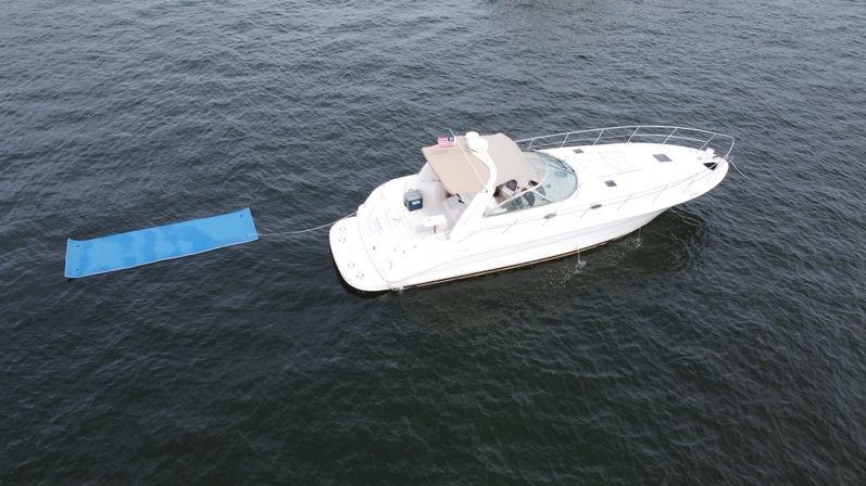 Aerial view of a sleek white motor yacht floating in open water, tethered to a bright blue floating mat off the stern.