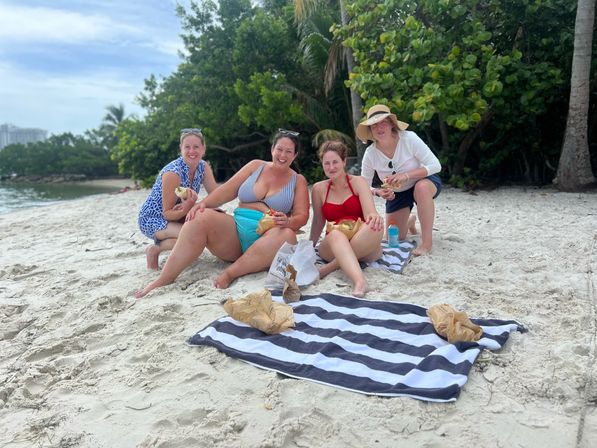 Four friends laughing and enjoying coconuts during a seaside picnic on a white-sand tropical beach with palm trees and calm ocean
