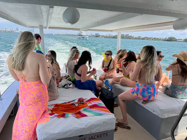 Group of women in colorful swimsuits enjoying a daytime boat party on turquoise waters with a distant Miami skyline, lounging under a shaded deck with towels and drinks.