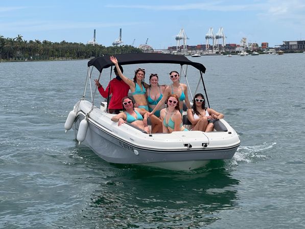 Group of friends in swimwear and heart-shaped sunglasses enjoying a sunny boat ride on a small motorboat with a black canopy in a palm-lined coastal harbor with cranes and boats in the background.