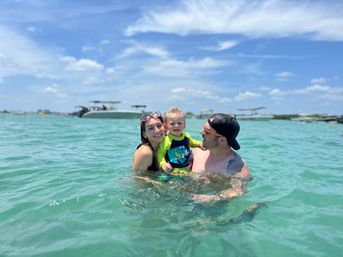 Smiling parents holding a toddler in shallow turquoise water near anchored boats under a sunny blue sky