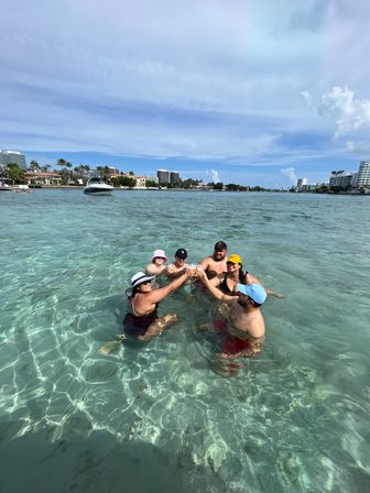 Six people toasting while standing in clear shallow turquoise water near anchored boats and waterfront buildings under a sunny blue sky