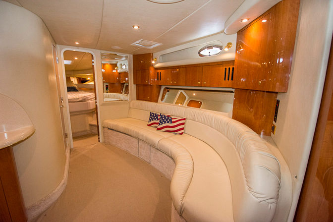 Cozy luxury yacht cabin interior with curved cream leather sofa, glossy cherry wood cabinets, porthole windows and two American-flag throw pillows.