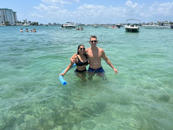 Smiling couple in swimsuits standing waist-deep in clear turquoise bay water holding a blue pool noodle, with anchored boats and a coastal skyline under a sunny blue sky.
