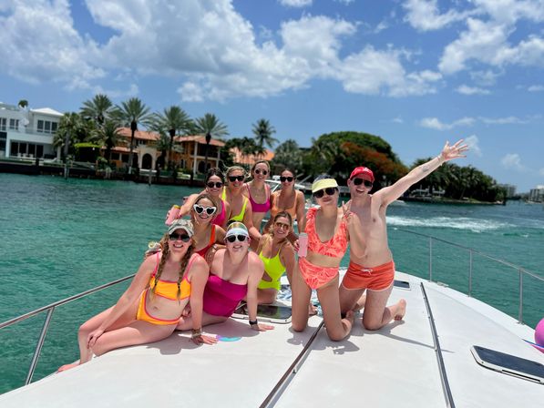 Group of friends in colorful swimsuits posing and smiling on a white yacht in turquoise coastal waters with palm-fringed waterfront homes under a sunny blue sky.