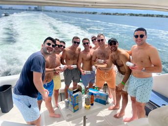 Nine friends on a motorboat party, shirtless in swim trunks and sunglasses, smiling and holding canned drinks on a sunny day with blue water, a trailing wake, and a distant shoreline.