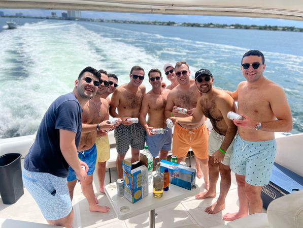 Nine friends on a motorboat party, shirtless in swim trunks and sunglasses, smiling and holding canned drinks on a sunny day with blue water, a trailing wake, and a distant shoreline.