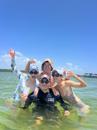Four smiling friends in shallow coastal water on a sunny summer day, wearing swimsuits, hats and sunglasses and toasting with plastic cups, boats anchored in the background.