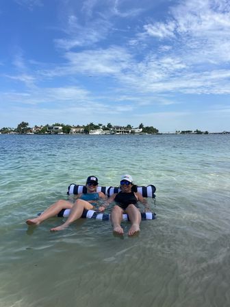 Two people in caps and sunglasses relaxing on a black-and-white striped float in shallow clear turquoise water near a palm-lined waterfront with houses under a bright blue sky.