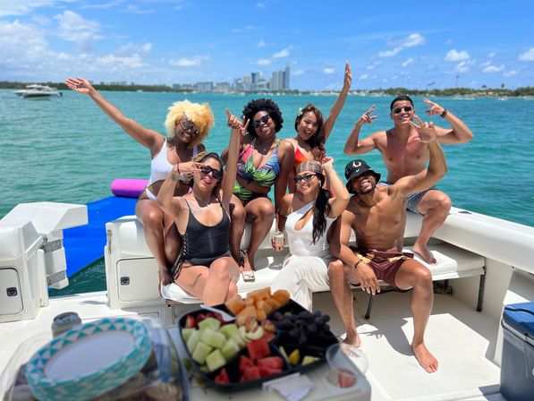 Seven friends in swimsuits cheering and posing on a sunny boat ride over turquoise water with the Miami skyline in the background and a fruit platter in the foreground