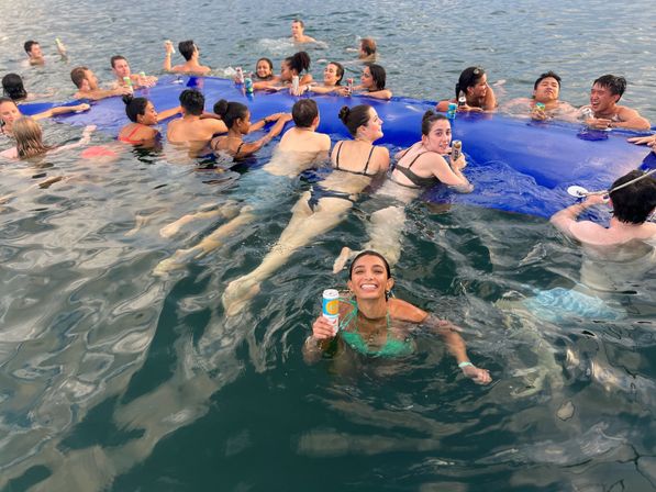 Smiling young adults at a waterfront summer swim party holding canned drinks and lounging on a large blue inflatable mat