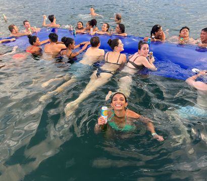 Smiling young adults at a waterfront summer swim party holding canned drinks and lounging on a large blue inflatable mat