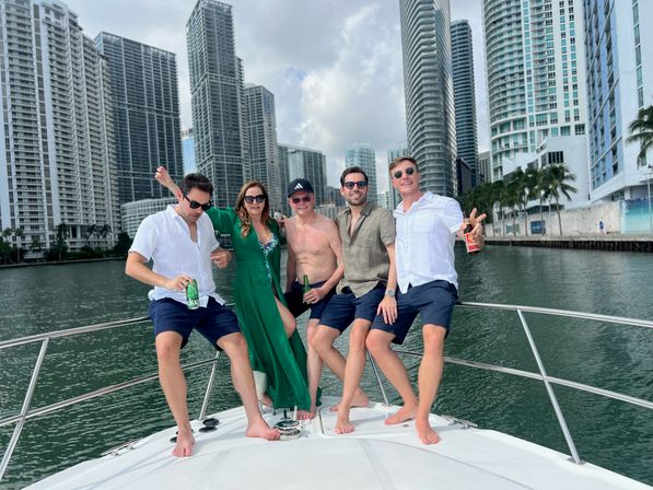 Five people posing barefoot on a yacht bow—men in casual shirts and shorts and a woman in a green dress—holding drinks and smiling with the Miami downtown high-rise waterfront skyline behind them on a sunny day.