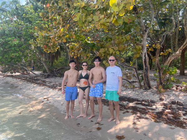Group of four people in swimwear posing on a sunny tropical beach vacation, standing on a sandy shoreline beneath sea grape trees with clear shallow water