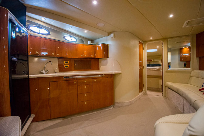 Luxury yacht cabin interior with glossy cherrywood galley cabinets, stainless sink and oval porthole windows, cream leather seating and carpeted floor leading to a cozy berth
