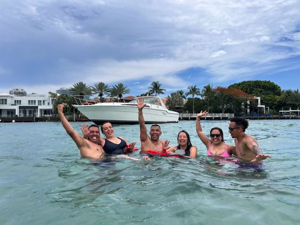Six people in swimsuits laughing and waving while floating in turquoise coastal water near a white yacht and palm-lined waterfront under a partly cloudy sky