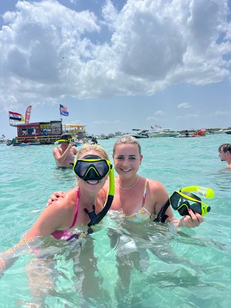 Two women in snorkeling masks wading in clear turquoise water near boats and a floating food bar, smiling under a sunny blue sky with puffy clouds — lively beach boating scene.
