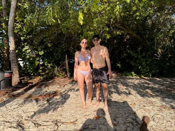 Two people posing on a sun-dappled tropical sandy beach beneath palm trees and dense green foliage — one in a lavender bikini and sunglasses, the other in patterned swim trunks and a cap.