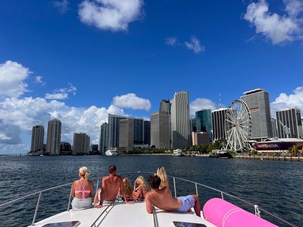 Group of people in swimwear relaxing on a boat bow cruising toward the Miami downtown skyline with a waterfront Ferris wheel and bright blue sky