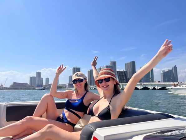Two friends in swimsuits and sunglasses cheering on a boat in Biscayne Bay with the Miami skyline and bridge under a bright blue sky