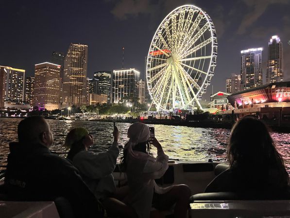 Silhouetted people on a boat photographing a giant illuminated Ferris wheel beside a downtown waterfront skyline at night, city lights reflecting on rippling water.