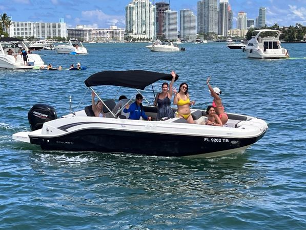 Friends waving and relaxing on a motorboat with a black canopy in a sunny urban bay, surrounded by other boats and a high-rise waterfront skyline.