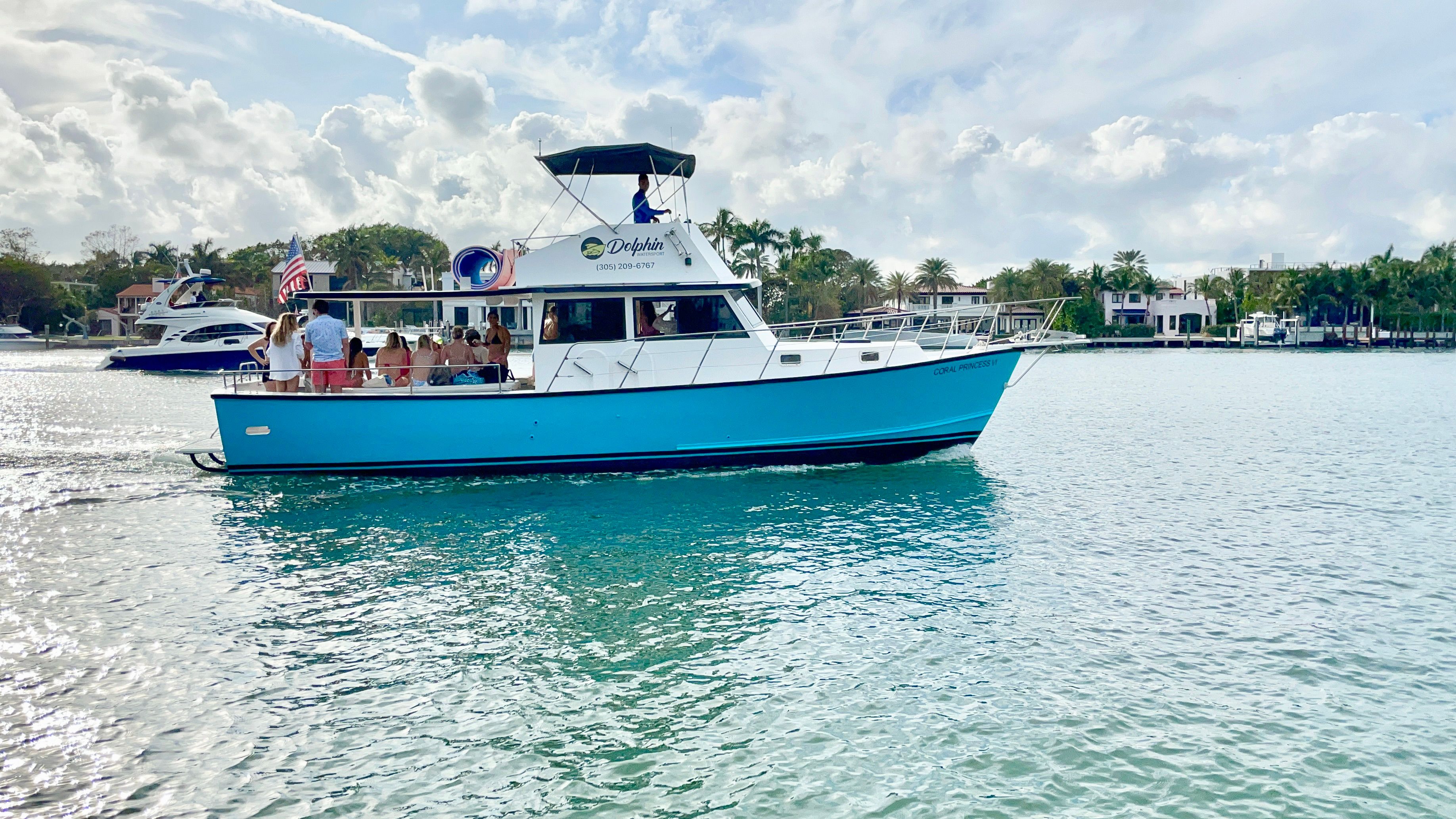 Bright blue motor yacht with passengers cruising a palm-lined coastal bay, sparkling turquoise water and waterfront homes under a sunny, partly cloudy sky.
