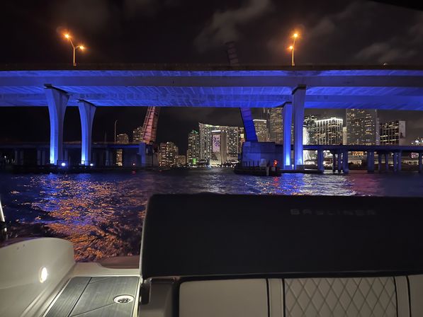 Boat-seat view under a blue-lit bridge framing a glittering downtown skyline at night, city lights shimmering on rippling water.
