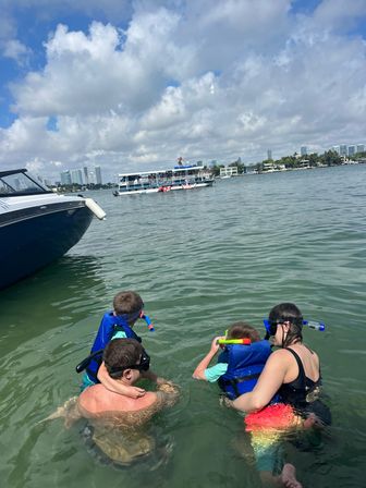 Family snorkeling in a sunny urban bay — kids in blue life jackets and snorkels beside a boat, with a tour boat and city skyline on the horizon
