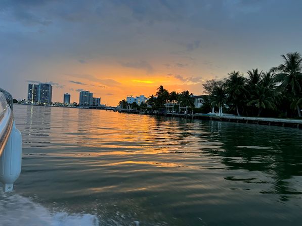 Boat-side view of a golden sunset over a palm-lined waterfront and distant condo skyline, reflections rippling across calm bay water.