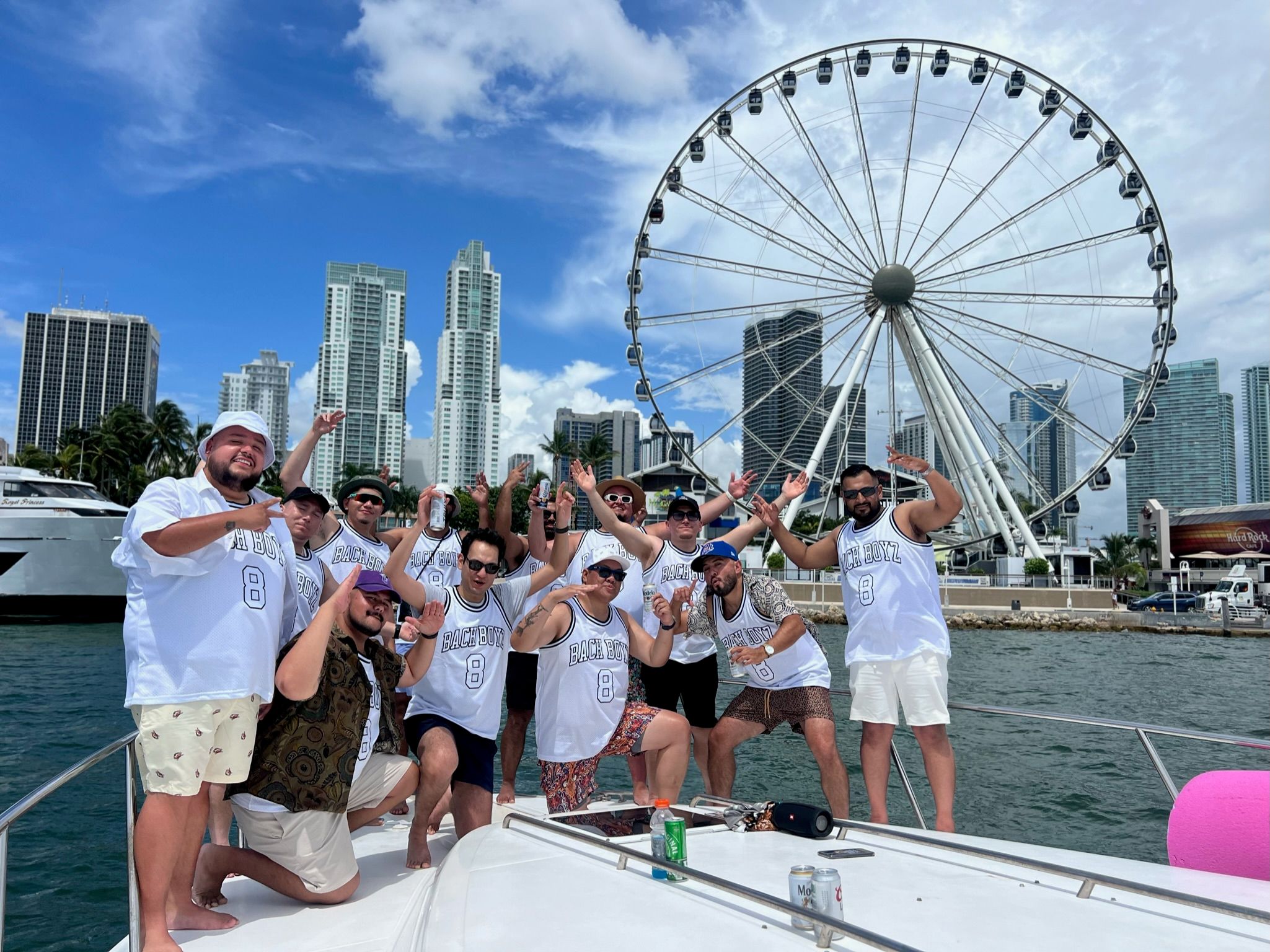 Group of friends in matching "Bach Boyz" jerseys posing and celebrating on a white yacht with a waterfront city skyline and large Ferris wheel under a bright blue sky.