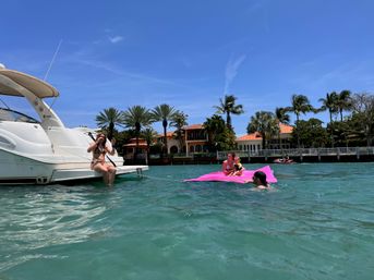 Sun-soaked boating scene on clear turquoise water by palm-lined tropical waterfront mansions — woman on a white yacht swim platform and a family lounging on a bright pink float under a vivid blue sky.