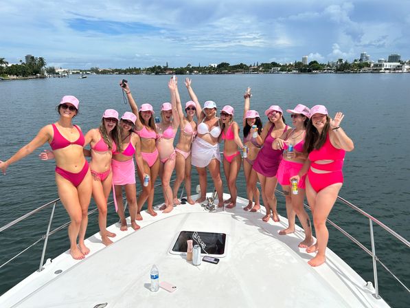 Friends in matching pink swimsuits and pink caps celebrating on the bow of a white yacht, waving and holding canned drinks with a calm bay, palm-lined shoreline and city skyline under a sunny blue sky — lively yacht/boat party scene.
