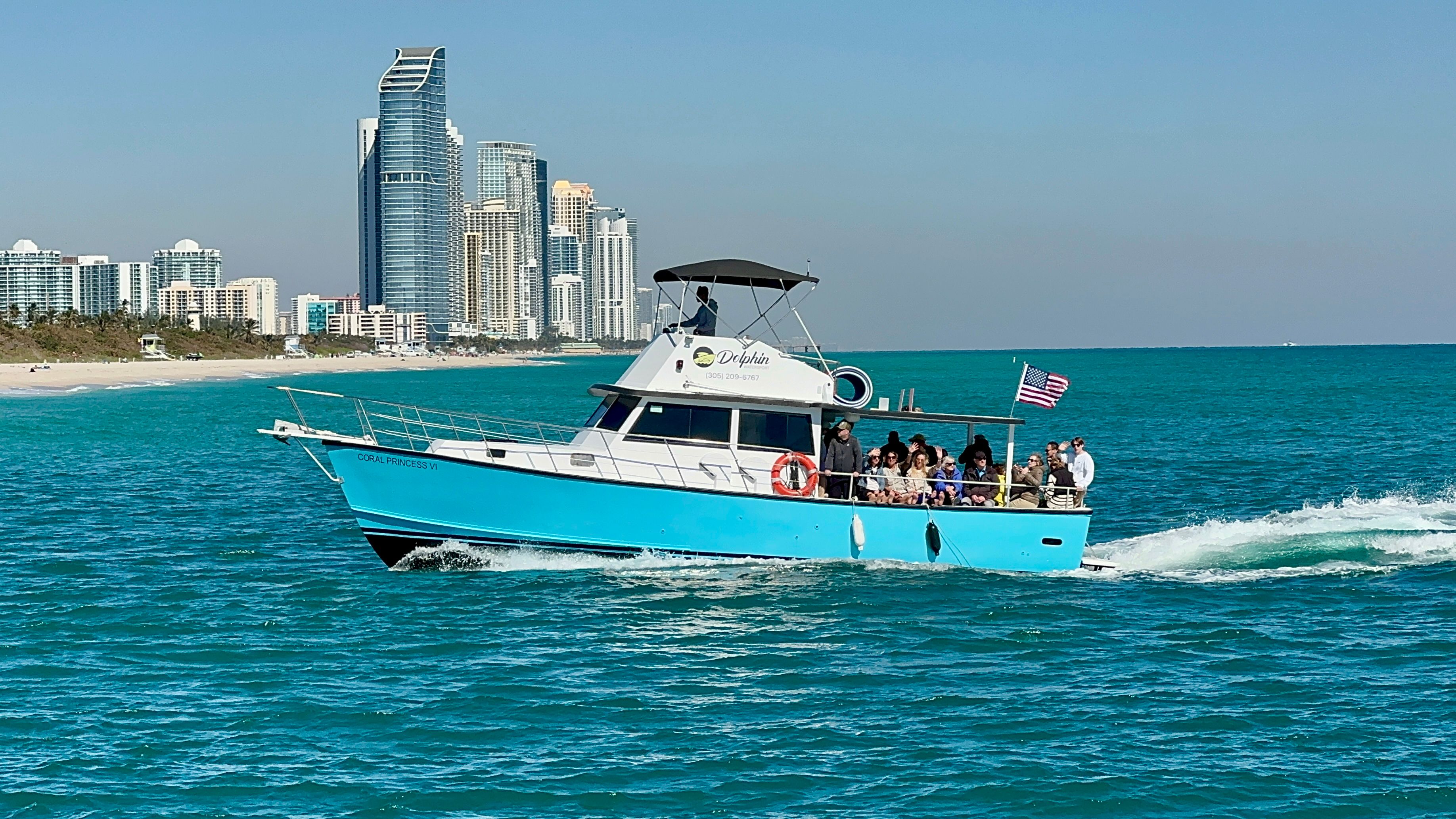 Bright turquoise tour boat with passengers and an American flag cruising on clear turquoise ocean near a sandy beach and high-rise coastal skyline on a sunny day