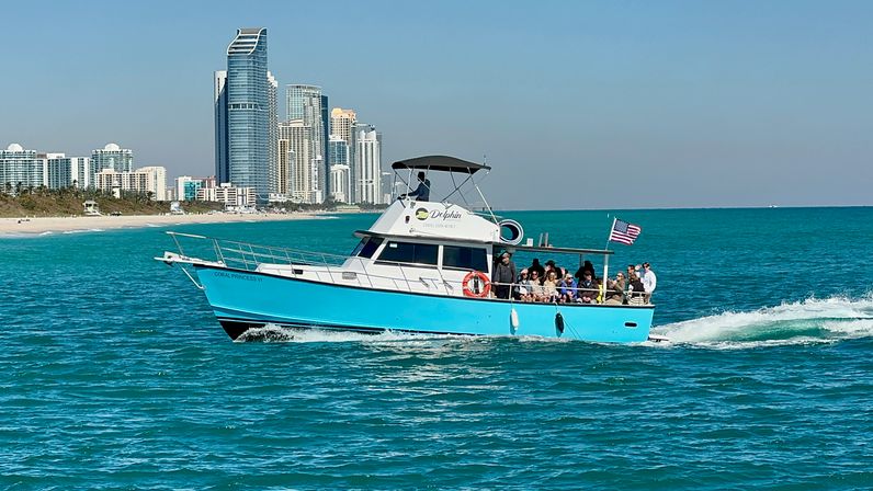 Bright turquoise tour boat with passengers and an American flag cruising on clear turquoise ocean near a sandy beach and high-rise coastal skyline on a sunny day