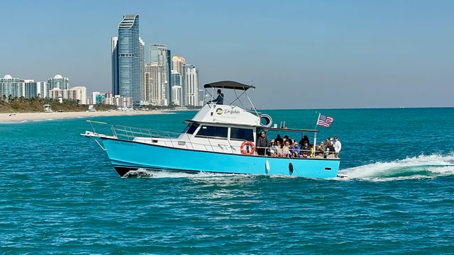 Bright turquoise tour boat with passengers and an American flag cruising on clear turquoise ocean near a sandy beach and high-rise coastal skyline on a sunny day