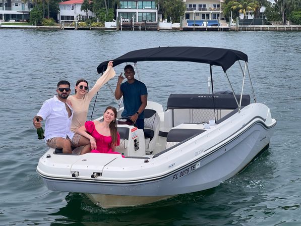Four friends enjoying a white motorboat with black canopy in a Florida waterfront bay, palm-lined homes in the background and one person holding a champagne bottle.