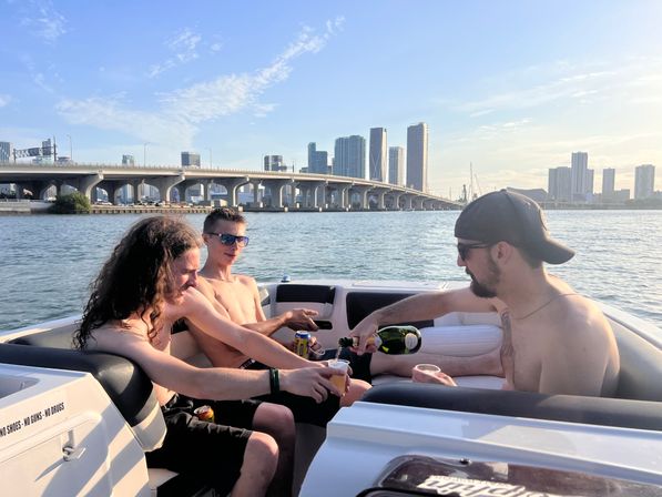 Three friends relaxing shirtless on a sunny motorboat near the Miami skyline and bridge, pouring drinks on calm bay water.