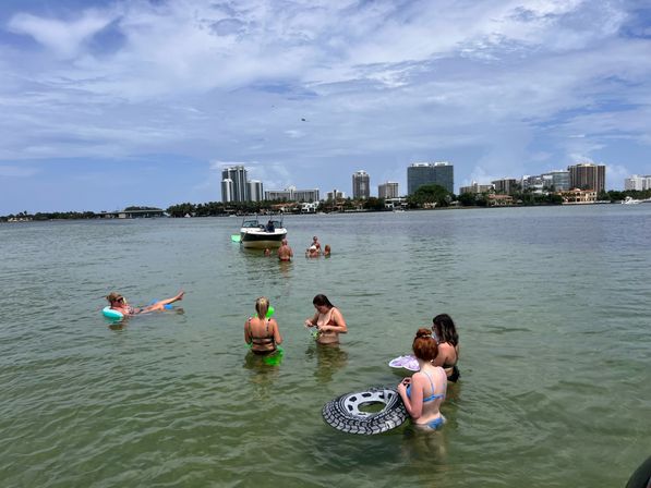 Group of people wading and floating in a shallow bay with a small motorboat and urban waterfront skyline under a partly cloudy sky