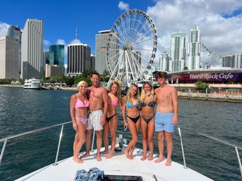 Group of six people in swimsuits posing on a yacht in sunny Miami Bay with the downtown skyline and a large waterfront Ferris wheel in the background.