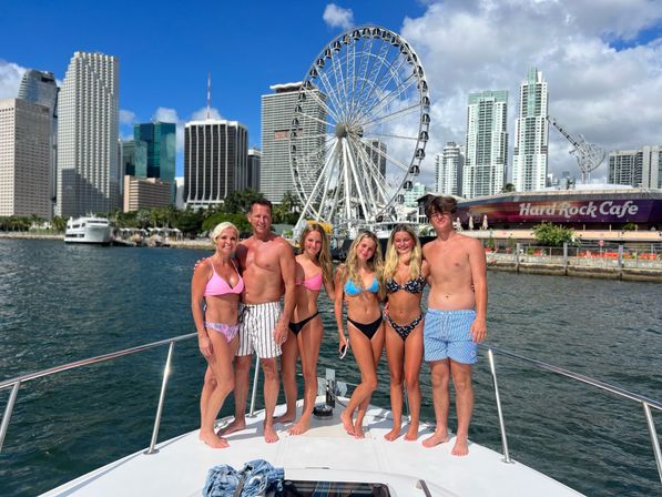 Group of six people in swimsuits posing on a yacht in sunny Miami Bay with the downtown skyline and a large waterfront Ferris wheel in the background.